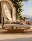 A luxury outdoor patio furniture set with a beige sofa, two matching armchairs, and a wooden coffee table, arranged on a woven rug. A woman is lounging on the sofa, and the background features a Mediterranean-style building with an archway, draped curtains, and a scenic ocean view.