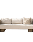 A modern outdoor sofa with thick cream cushions and chunky light wood arms and legs, set against a neutral beige wall with soft leaf shadows.